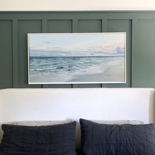 A framed horizontal landscape painting of an ocean beach hangs on a dark green paneled wall above a bed with a white upholstered headboard and dark gray bedding.